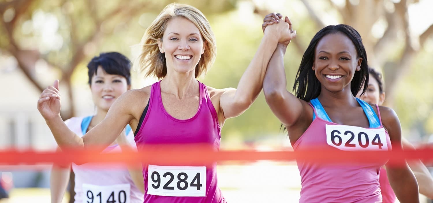 Female runners crossing finish line with raised arms at a race event in Brooklyn.