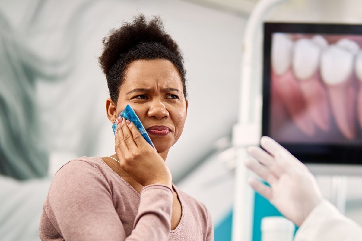 Woman experiencing tooth pain holds ice pack to cheek during dental visit in Brooklyn.