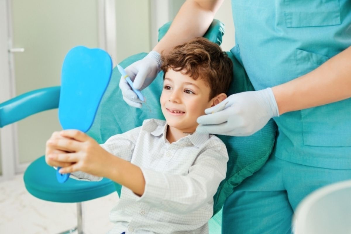 Young boy smiling in dental chair while pediatric dentist examines his teeth with mirror.